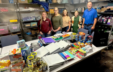 Staff from the HPFC Hays office stand behind a table in the First Call For Help of Ellis County office. The table holds a variety of school supplies and non-perishable food items.