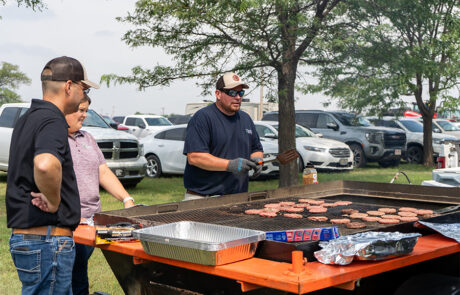 Two members of the HPFC team stand beside a large grill on a flatbed trailer. One employee flips burgers that are cooking.