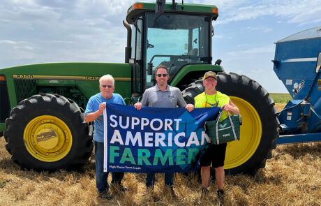HPFC VP Loan Officer, Wade Gantz, stands between two customers, holding a Support American Farmers flag. A green tractor makes up the background.