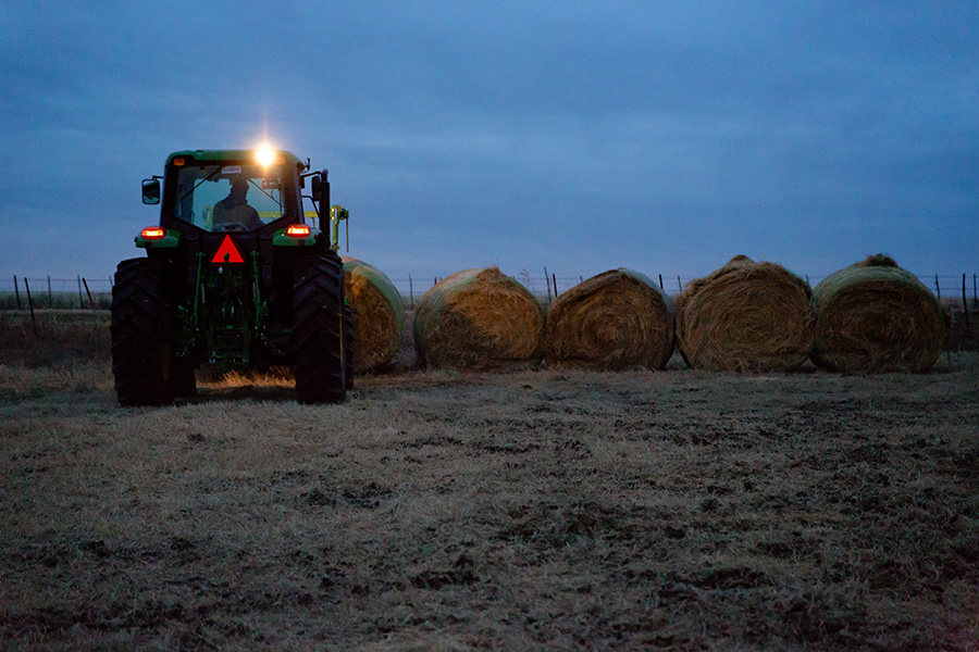 Farmer in a tractor lines up large, round hay bales as the sun sets over a cold evening.