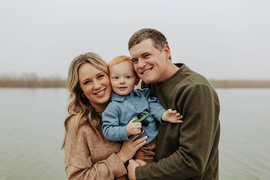 The McMillen family smiles as they stand in front of a lake on a cool autumn day.